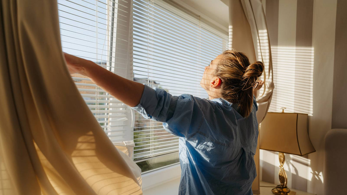 woman opening curtains to a sunny city view.