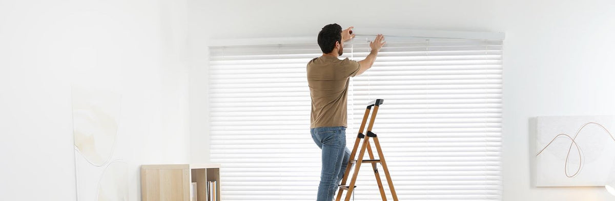 A man on a ladder installing faux wood blinds into a window.